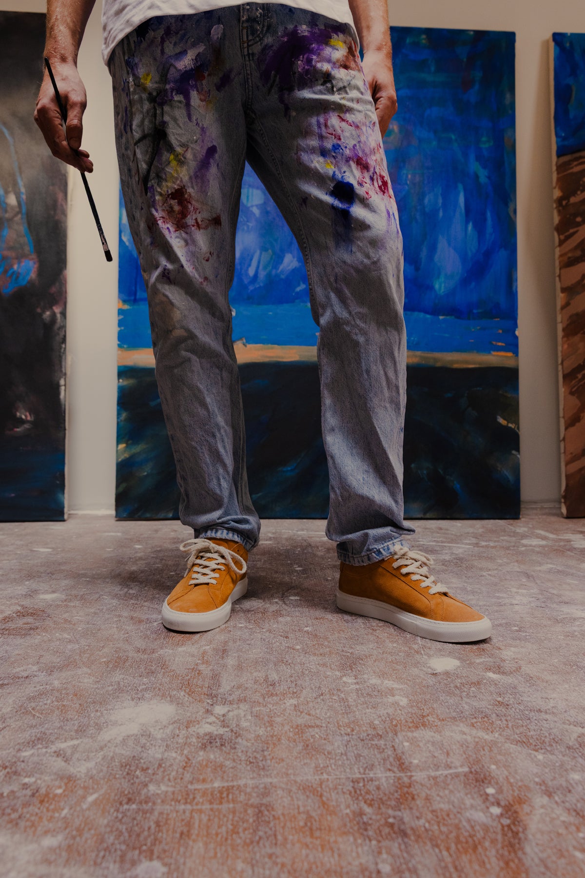 Person wearing paint-splattered pants standing in front of artworks.